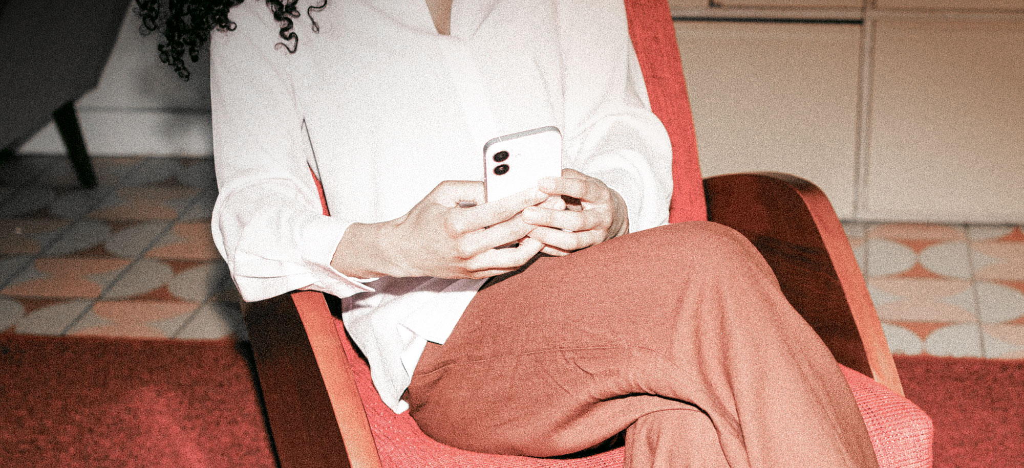 Person mit lockigem Haar sitzt in rotem Sessel und schaut auf ihr Smartphone.