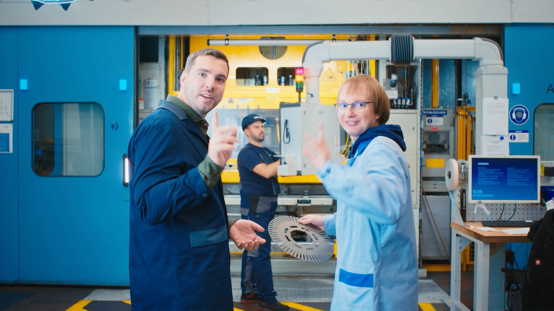 Zwei Mitarbeitende im Produktionsbereich winken in die Kamera, eine Person hält ein metallisches Bauteil.