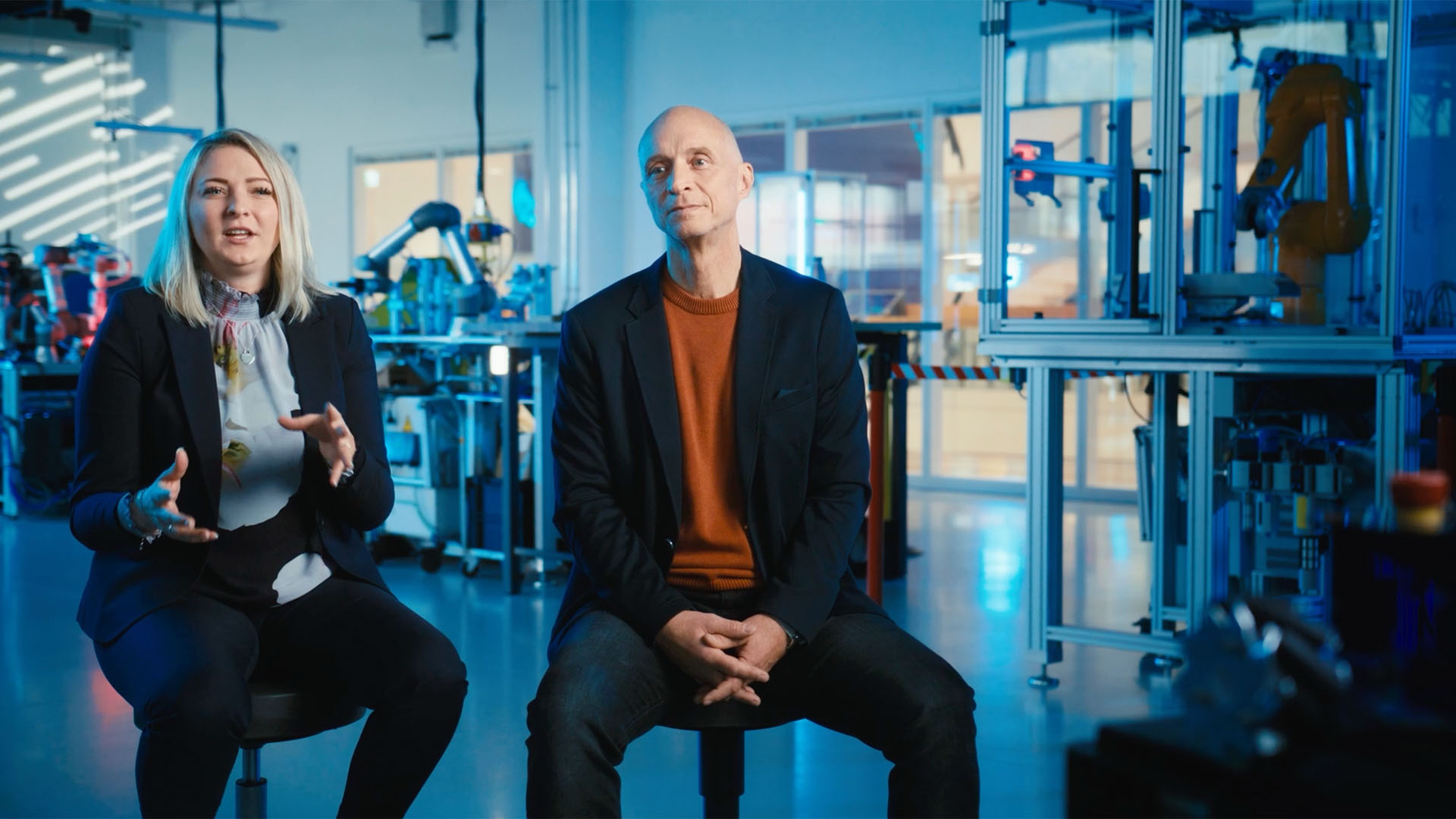 Zwei Personen in Business-Outfits sitzen in einem technischen Produktionsumfeld.