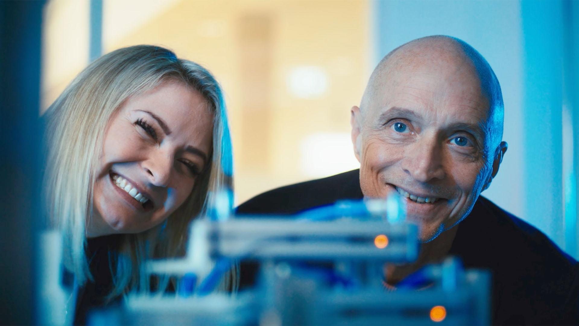 Zwei Personen lächeln in die Kamera, im Vordergrund unscharfes technisches Gerät.