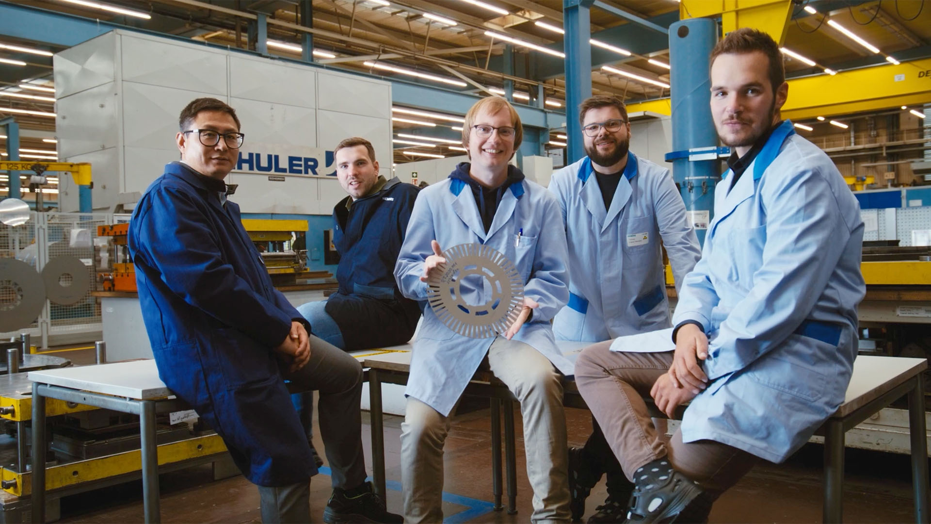 Personen in Arbeitskleidung in einem Produktionsumfeld, eine Person hält ein metallisches Bauteil.
