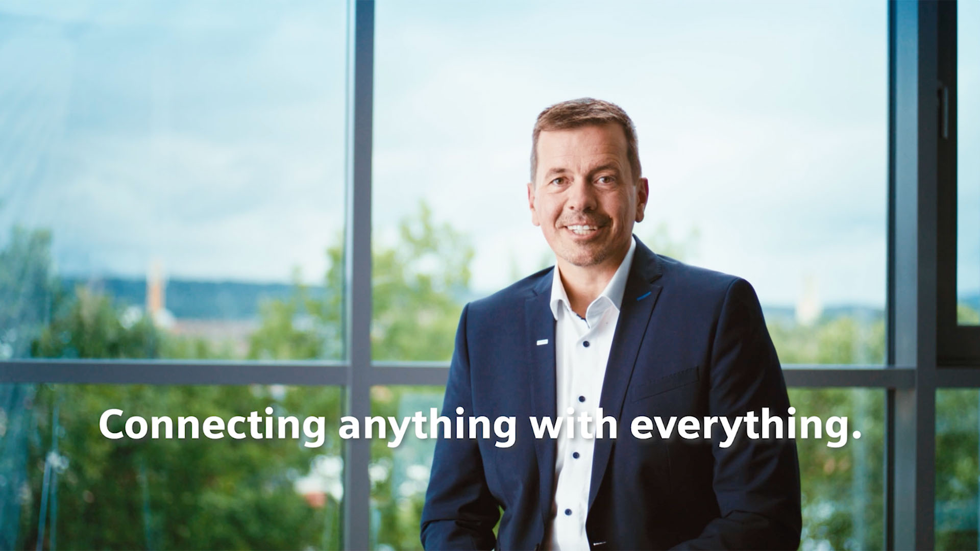 Porträt eines Mannes im Business-Outfit vor einer Fensterfront, mit eingeblendetem Claim „Connecting anything with everything“.
