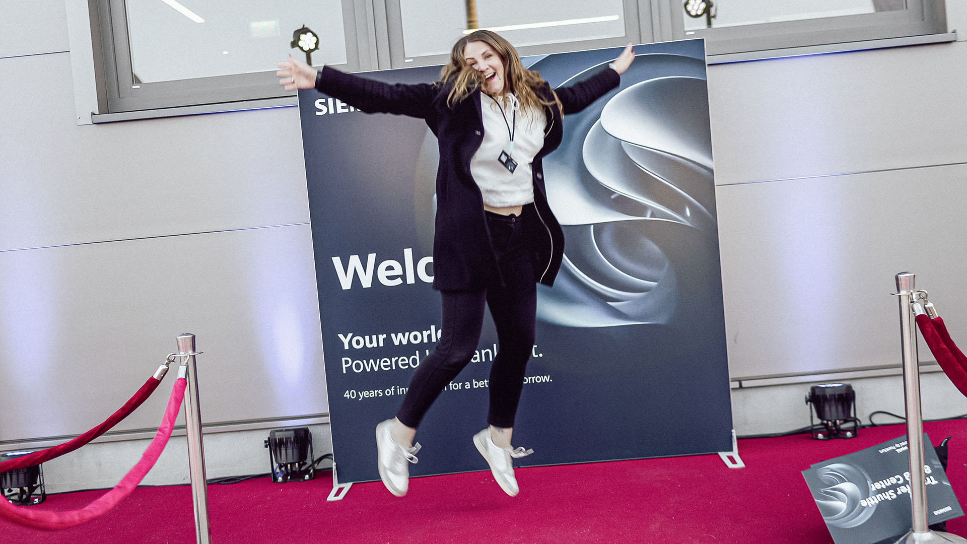 Eine Frau springt lächelnd vor einer „Welcome“-Wand mit rotem Teppich in die Luft.
