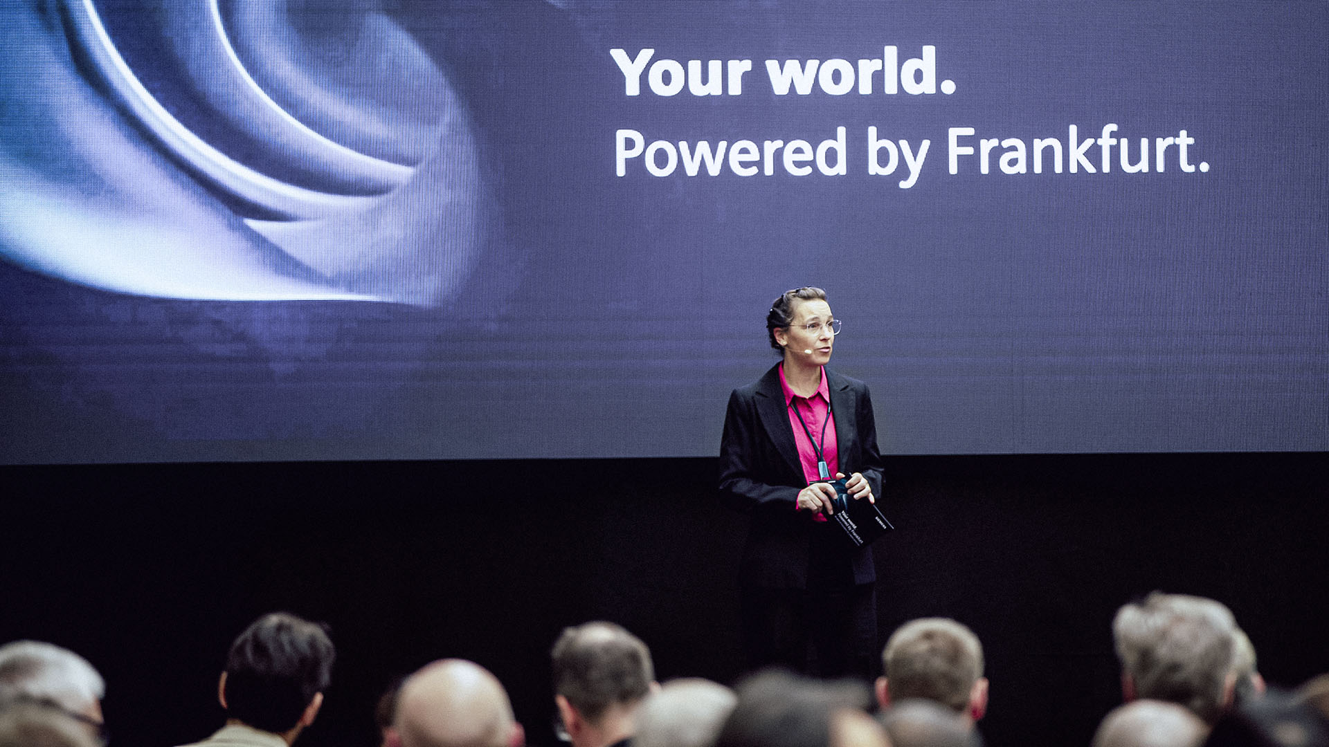 Eine Rednerin steht vor einem großen Bildschirm mit der Aufschrift „Your world. Powered by Frankfurt.“ vor einem Publikum.