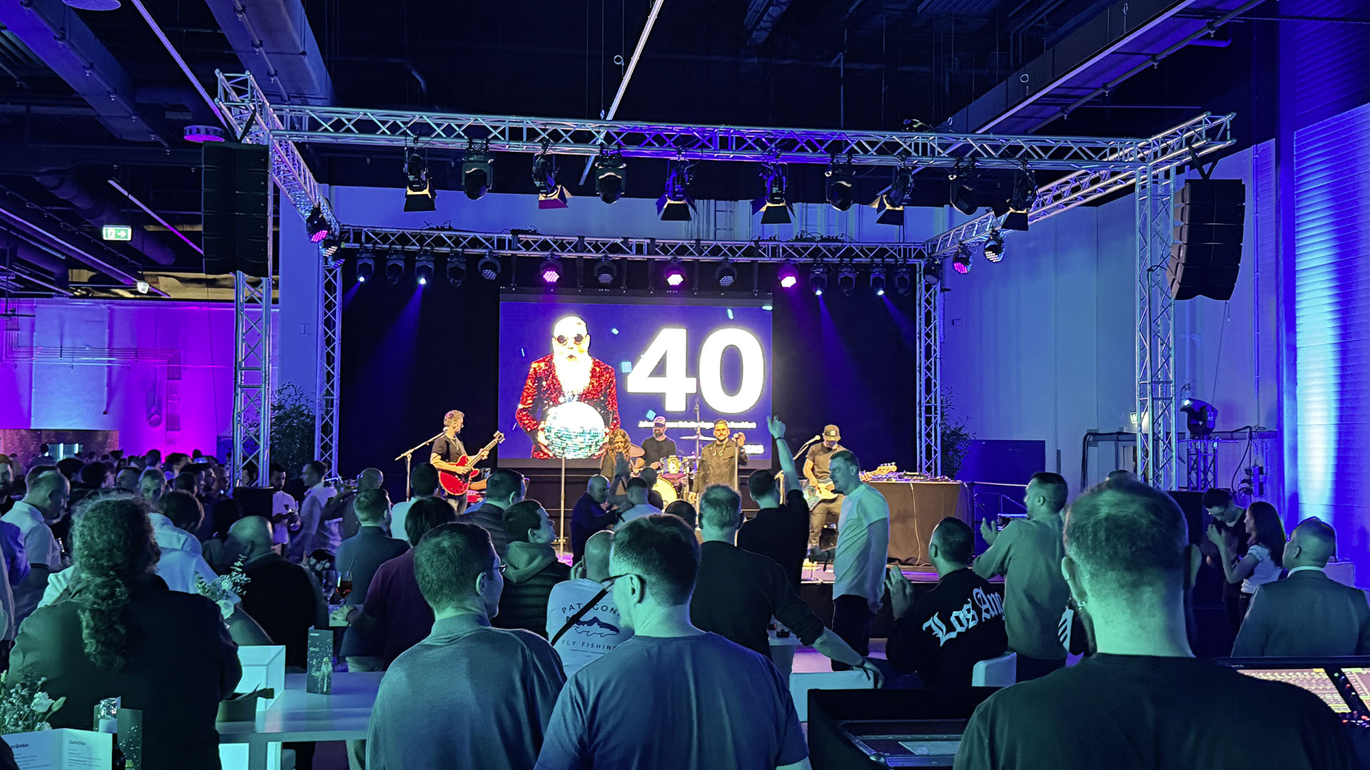 Eine große Menschenmenge schaut auf eine beleuchtete Bühne, auf der eine Band spielt und ein Bildschirm eine „40“-Jubiläumsgrafik zeigt.