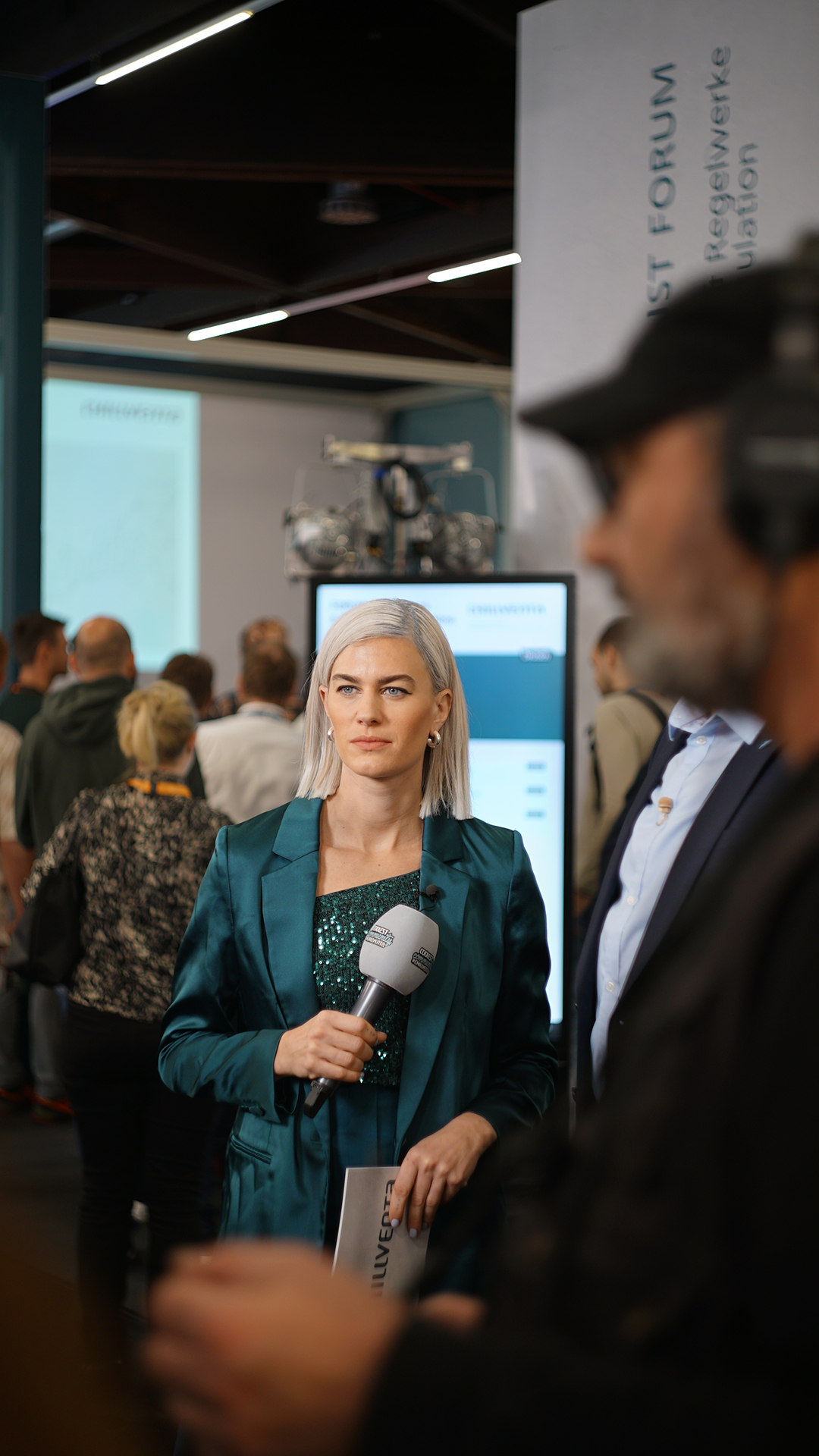 Frau mit Mikrofon in einer Messehalle, Menschen und Messestände im Hintergrund.