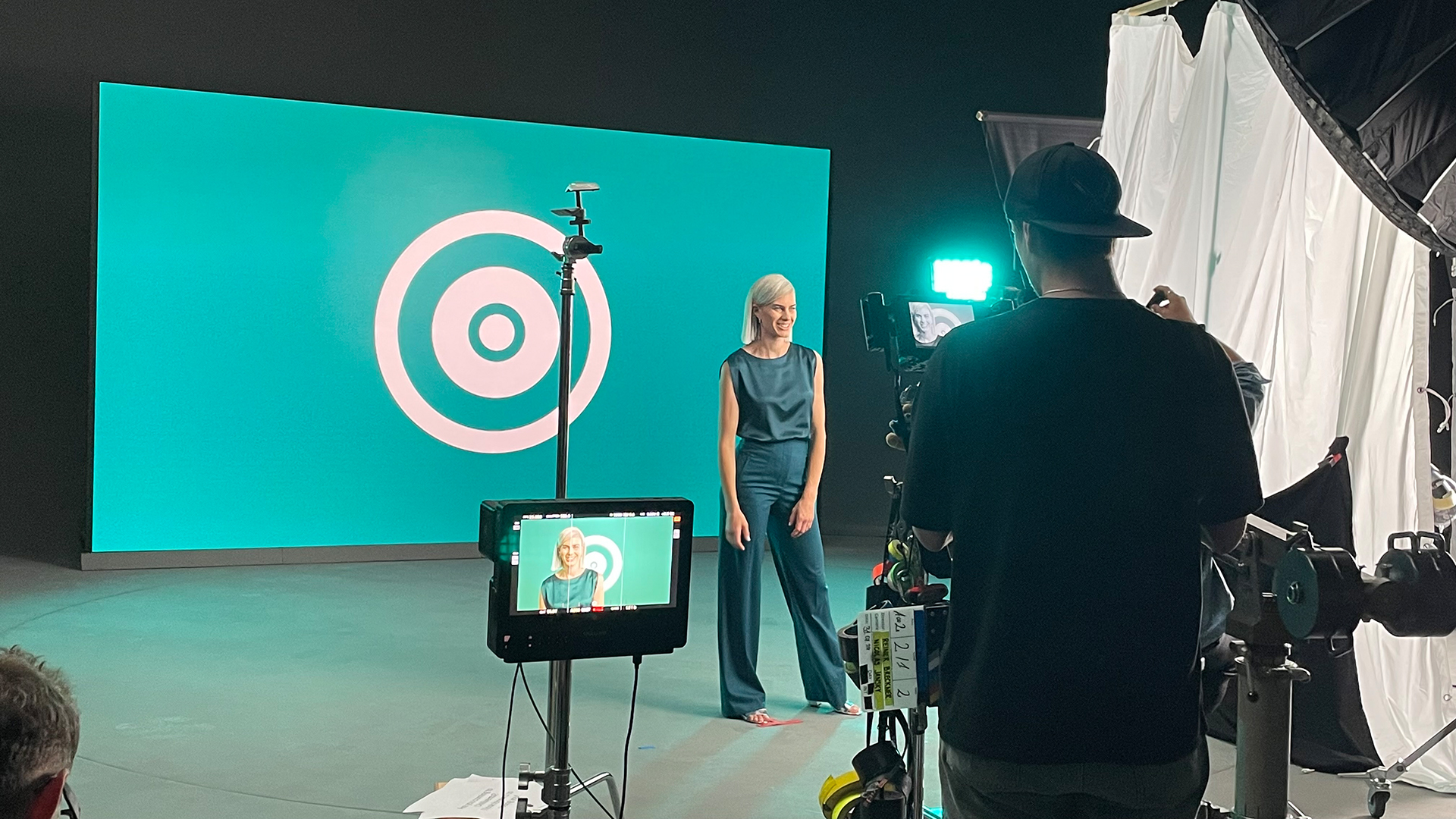Woman in front of a turquoise background during a video shoot, camera visible in the foreground.